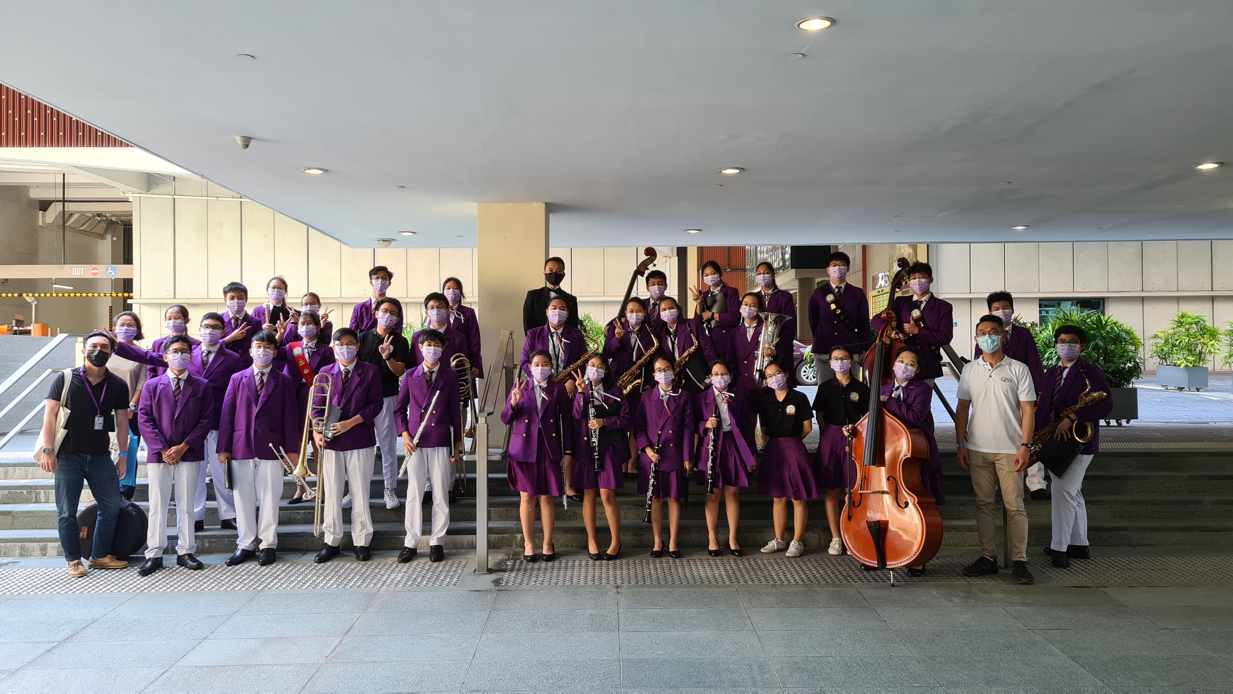 A group of students in purple uniforms wearing masks pose with orchestra instruments including a double bass and saxophones.